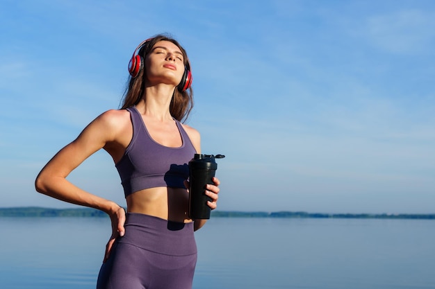 Uma jovem de agasalho e fones de ouvido está descansando do lado de fora depois de um treino Contra o fundo da água do mar clara