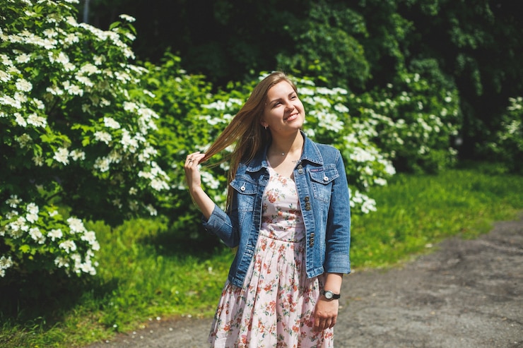 Foto uma jovem de aparência europeia com longos cabelos loiros, vestida com um vestido curto, está de pé contra o fundo de arbustos de flores brancas. dia ensolarado de primavera. beleza feminina natural