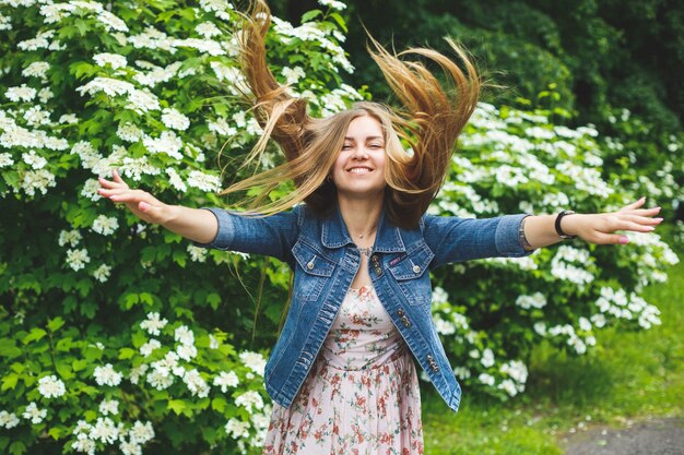 Foto uma jovem de aparência europeia com longos cabelos loiros, vestida com um vestido curto, está de pé contra o fundo de arbustos de flores brancas. dia ensolarado de primavera. beleza feminina natural