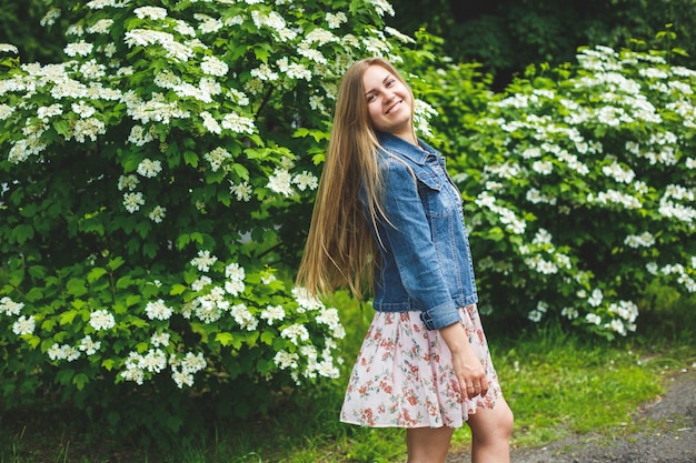 Uma jovem de aparência europeia com longos cabelos loiros, vestida com um vestido curto, está de pé contra o fundo de arbustos de flores brancas. Dia ensolarado de primavera. Beleza feminina natural