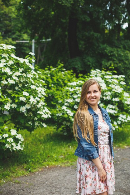 Uma jovem de aparência europeia com longos cabelos loiros, vestida com um vestido curto, está de pé contra o fundo de arbustos de flores brancas. Dia ensolarado de primavera. Beleza feminina natural