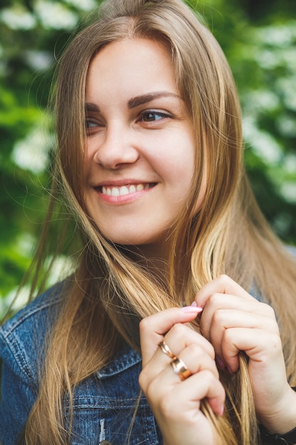 Uma jovem de aparência europeia com longos cabelos loiros, vestida com um vestido curto, está de pé contra o fundo de arbustos de flores brancas. dia ensolarado de primavera. beleza feminina natural