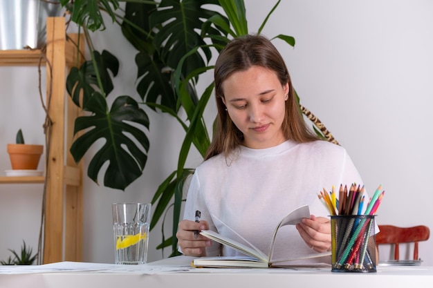 Uma jovem de aparência europeia faz lição de casa na universidade escolar A menina estuda diligentemente em casa em uma mesa branca Fotografia em tons claros no interior de uma casa