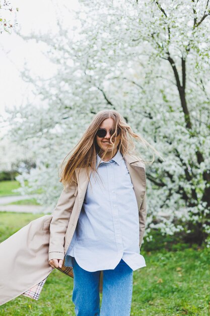 Uma jovem de cabelos loiros desfruta de um jardim de primavera em flor Viajar férias de primavera Estilo elegante Uma mulher de óculos escuros e um casaco bege atravessa um parque florido Foco de seleção