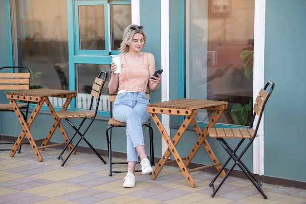 Uma jovem de calças jeans senta-se com uma caneca de café e um telefone perto de um café no verão