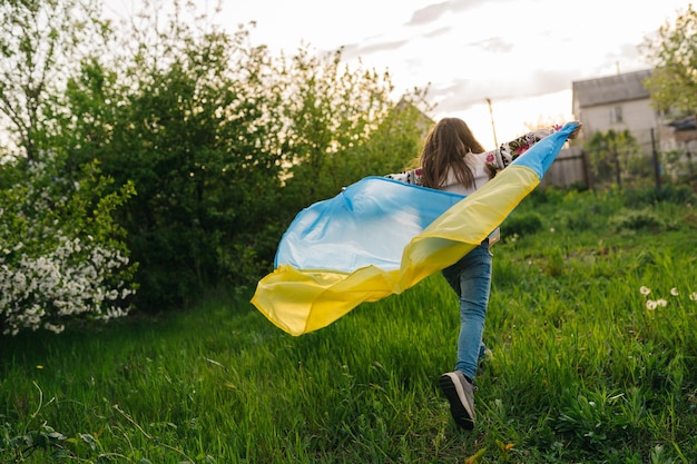 Uma jovem de camisa bordada corre com a bandeira ucraniana Foto de uma menina correndo com uma bandeira ucraniana usada como capa