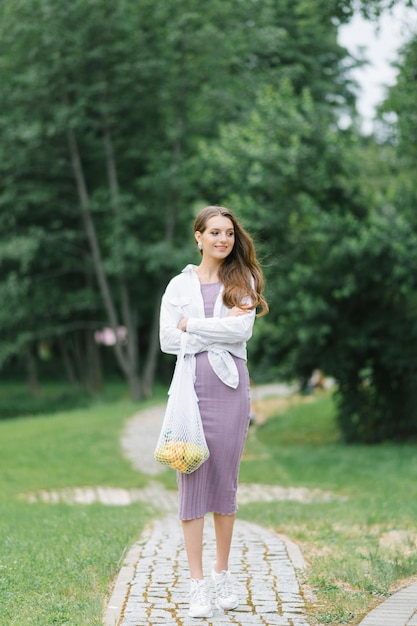 Uma jovem de camisa branca, um vestido lilás com um saco de frutas ecologicamente correto