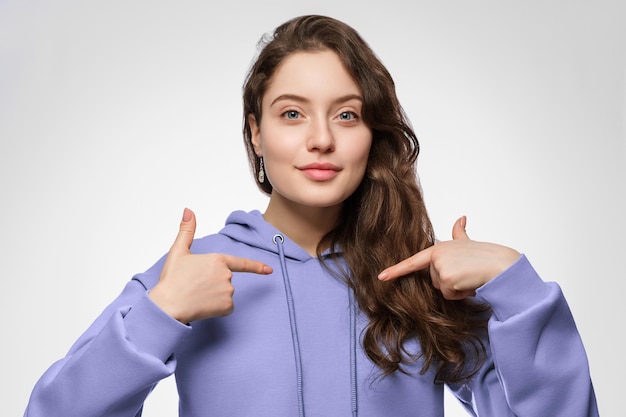 Uma jovem de capuz aponta para si mesma com os dedos indicadores. Cabelo comprido e olhos azuis.