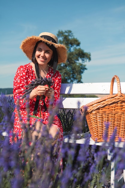 Uma jovem de chapéu de palha senta-se em um banco em um campo de lavanda e tece uma coroa de flores