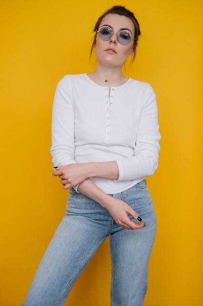 Uma jovem de moletom branco e jeans azul fica no estúdio contra um fundo amarelo