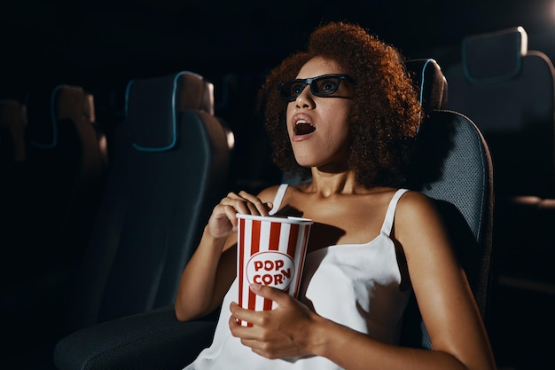 Uma jovem de óculos 3d está assistindo a um filme com entusiasmo pipoca nas mãos