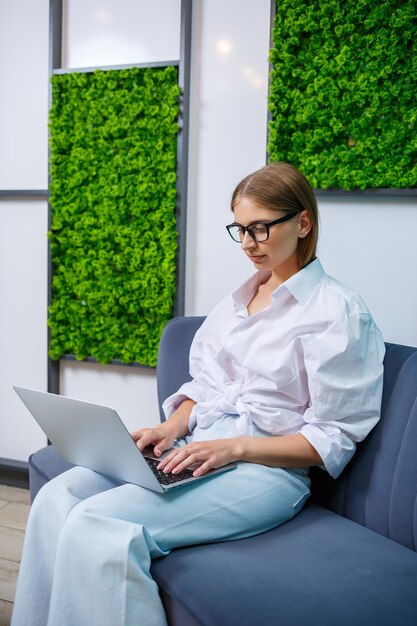 Uma jovem de óculos e uma camisa de algodão branca se senta em um sofá e trabalha online em um laptop trabalho remoto feminino freelancer trabalhando em casa