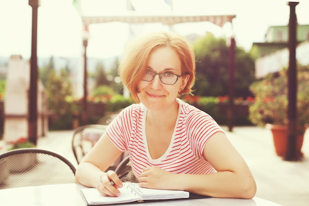 Uma jovem de óculos se senta em uma mesa e faz anotações em um caderno, um clima leve e quente de verão