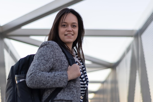 Uma jovem de pé segurando sua mochila no ombro olhando para a câmera com um sorriso