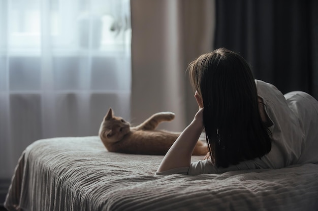 uma jovem de pijama está deitada na cama perto da janela ao lado de seu gato doméstico