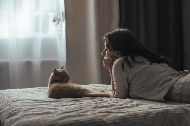 uma jovem de pijama está deitada na cama perto da janela ao lado de seu gato doméstico