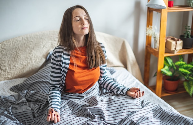 Uma jovem de pijama está sentada na cama de manhã na posição de lótus e meditando