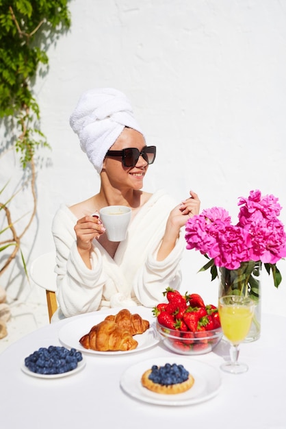Uma jovem de roupão de banho e toalha na cabeça aproveita suas férias no resort toma café da manhã com croissant e morangos