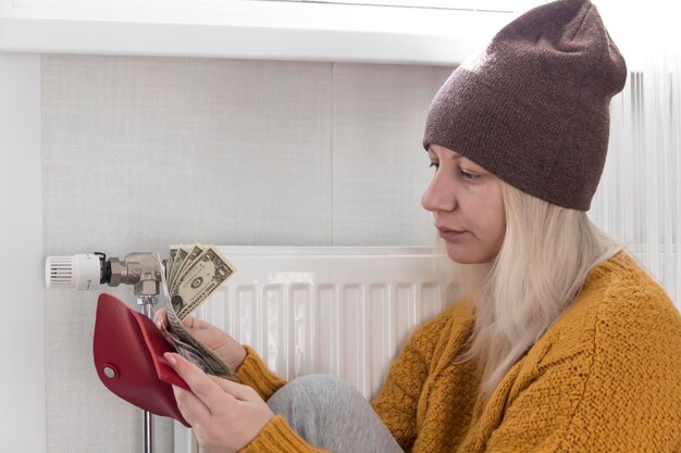 Uma jovem de suéter amarelo e chapéu marrom está sentada no chão contando dinheiro e pensando em como pagar contas e impostos perto de um aquecedor com termostato