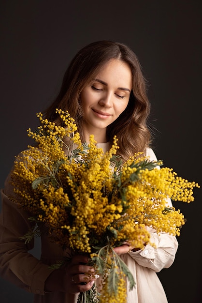 Uma jovem de vestido bege segura um buquê de mimosa em suas mãos e olha para ele