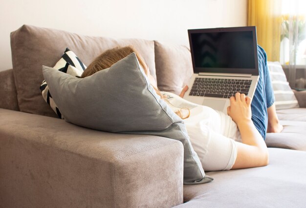 Uma jovem deita-se no sofá da sala e trabalha usando um laptop em casa.