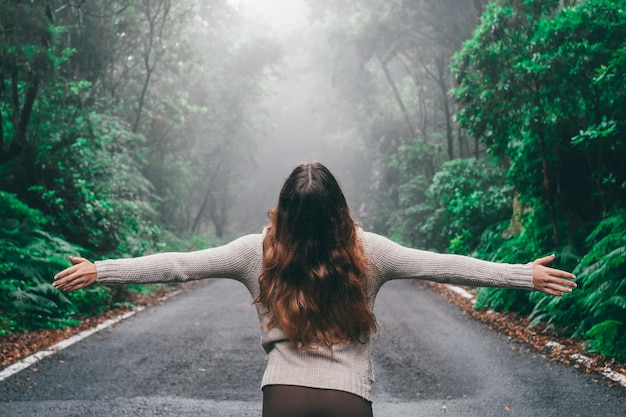 Uma jovem desfrutando e descobrindo a floresta sozinha andando no meio da estrada com árvores verdes ao redor dela garota abrindo os braços e olhando para cima se sentindo livre conceito de liberdade