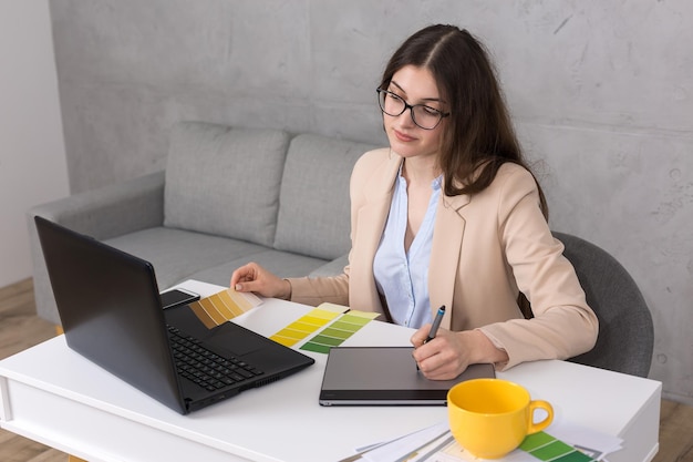 Uma jovem designer sentada em uma mesa desenha em um tablet Faz cálculos de negócios no computador