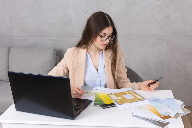 Uma jovem designer sentada em uma mesa desenha em um tablet faz cálculos de negócios no computador