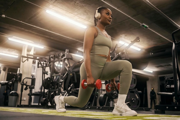 Uma jovem desportista negra em forma a praticar lunges com halteres e a ouvir música.