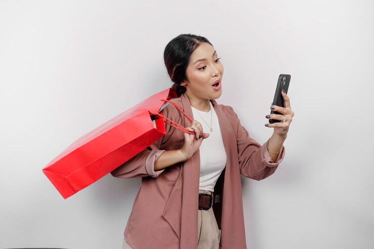 Foto uma jovem e bela funcionária asiática surpresa vestindo um cardigan está carregando uma bolsa de compras on-line e seu smartphone isolado em fundo branco