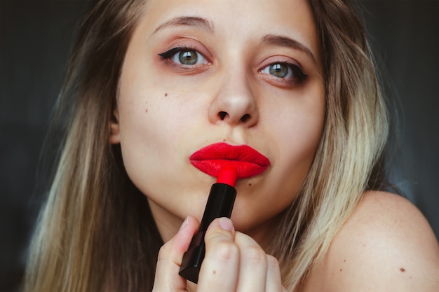 Uma jovem e bela loira caucasiana com manchas no rosto pinta os lábios com batom vermelho em um fundo escuro.