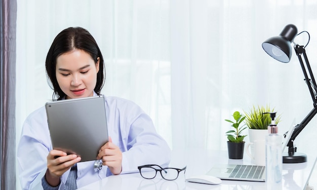 Uma jovem e bela médica asiática no computador portátil frontal da mesa, ela segurando um tablet digital inteligente moderno disponível durante a quarentena, doença coronavírus ou COVID-19 no escritório do hospital