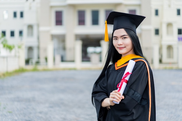 Uma jovem e bela mulher asiática, graduada em vestido de formatura e papelão com um diploma universitário, está de pé em frente ao prédio da universidade após participar da formatura da faculdade