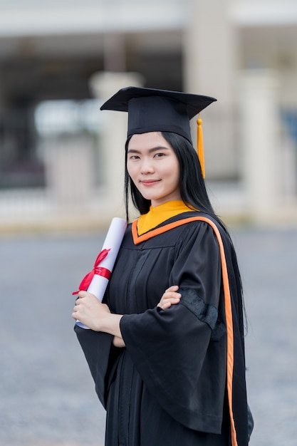 Uma jovem e bela mulher asiática, graduada em vestido de formatura e papelão com um diploma universitário, está de pé em frente ao prédio da universidade após participar da formatura da faculdade