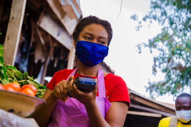 Uma jovem e bela mulher do mercado africano usando máscara facial para se prevenir da sociedade segurando uma máquina pos.