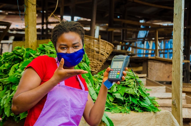 Uma jovem e bela mulher do mercado africano usando máscara facial para se prevenir da sociedade segurando uma máquina pos.