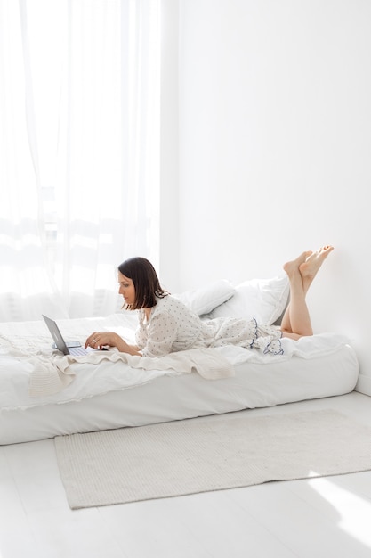 Uma jovem e bela mulher europeia deitada em uma cama em um quarto branco com um laptop