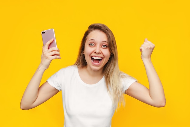 Uma jovem e bonita caucasiana impressionada, animada e sorridente, mulher loira com uma camiseta branca e um telefone celular na mão está feliz com a notícia ou com a vitória na loteria isolada em uma parede de cor amarela brilhante