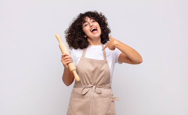 Uma jovem e bonita chef se sentindo feliz, surpresa e orgulhosa, apontando para si mesma com um olhar animado e surpreso