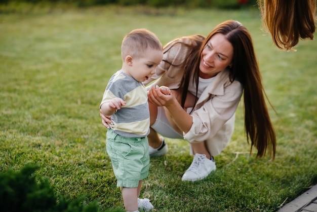 Uma jovem e bonita mãe ajuda e ensina seu filho a dar os primeiros passos durante o pôr do sol no parque na grama.