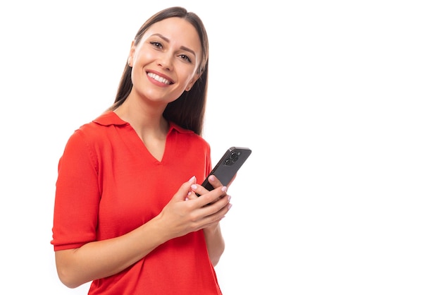 Uma jovem e bonita mulher europeia com cabelo preto está vestida com uma camiseta vermelha com um smartphone em um