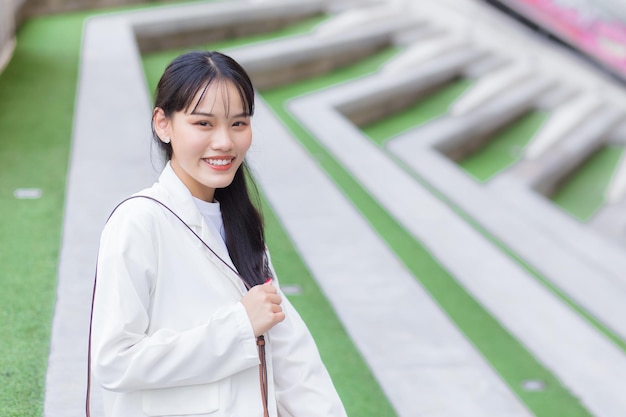Uma jovem e confiante mulher trabalhadora asiática que usa uma camisa branca e uma bolsa de ombro sorri alegremente