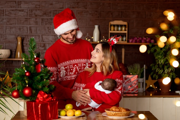 Uma jovem e feliz família, mãe, pai e bebê em suéteres vermelhos na cozinha com uma árvore de natal em casa celebram o ano novo ou o natal regozijando-se com o nascimento de uma criança