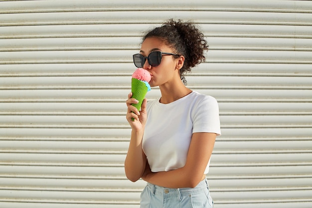 Uma jovem e linda garota afro-americana em uma camiseta branca e jeans claro come sorvete contra uma parede leve em um dia ensolarado