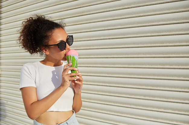 Uma jovem e linda garota afro-americana em uma camiseta branca e jeans claro come sorvete contra uma parede leve em um dia ensolarado