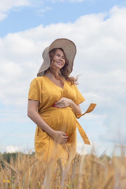 Uma jovem e linda mulher grávida com um vestido e chapéu amarelo caminha por um campo de trigo laranja em um dia ensolarado de verão