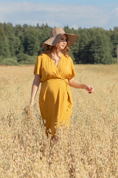 Uma jovem e linda mulher grávida com um vestido e chapéu amarelo caminha por um campo de trigo laranja em um dia ensolarado de verão