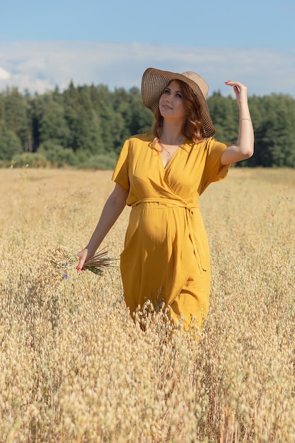 Uma jovem e linda mulher grávida com um vestido e chapéu amarelo caminha por um campo de trigo laranja em um dia ensolarado de verão