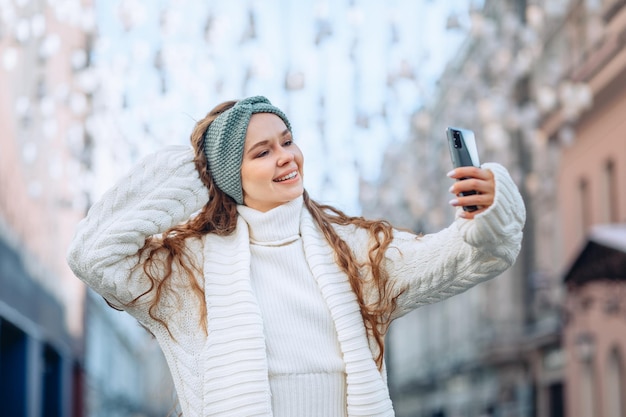 Uma jovem elegante e deslumbrante, com cabelos longos e encaracolados, vestida com um terno branco de malha quente tira uma selfie na cidade. uma mão levantada e um sorriso brilhante. o conceito de uma selfie de alta qualidade.