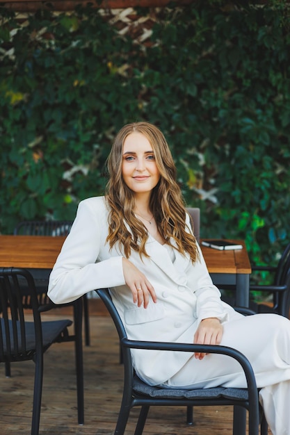 Uma jovem elegante em um terno branco clássico com um sorriso no rosto está no terraço de um café de verão